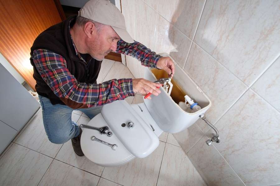 A mature male technician wearing a plaid shirt and cap uses a small tool to repair the internal mechanism of a running toilet.