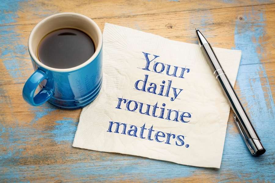 A blue coffee mug and a pen next to a napkin that reads your daily routine matters on a wooden table.