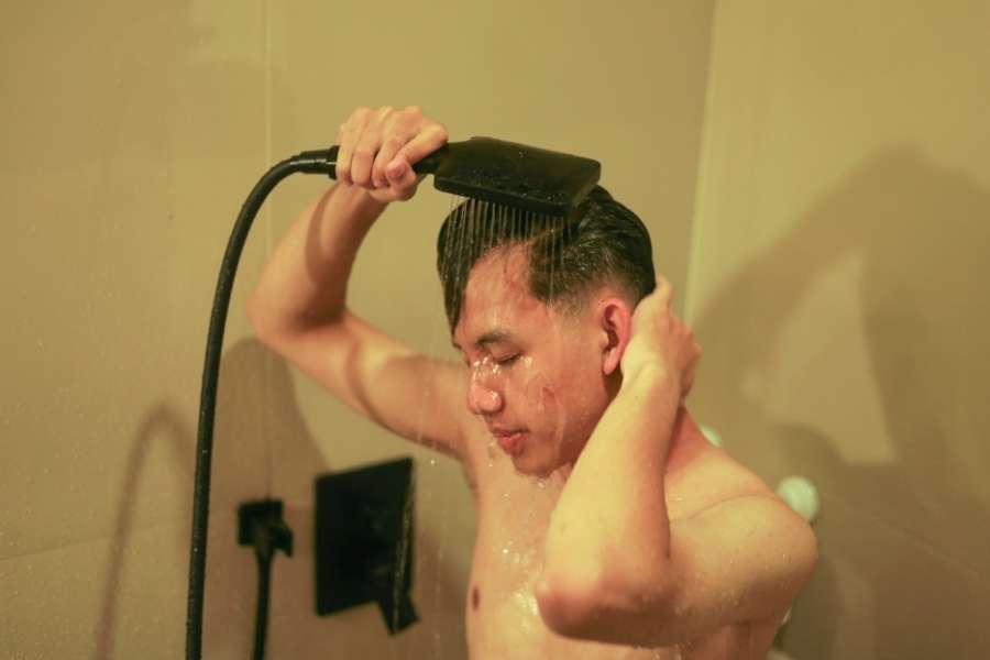 A young man washes his hair while holding a black handheld showerhead.